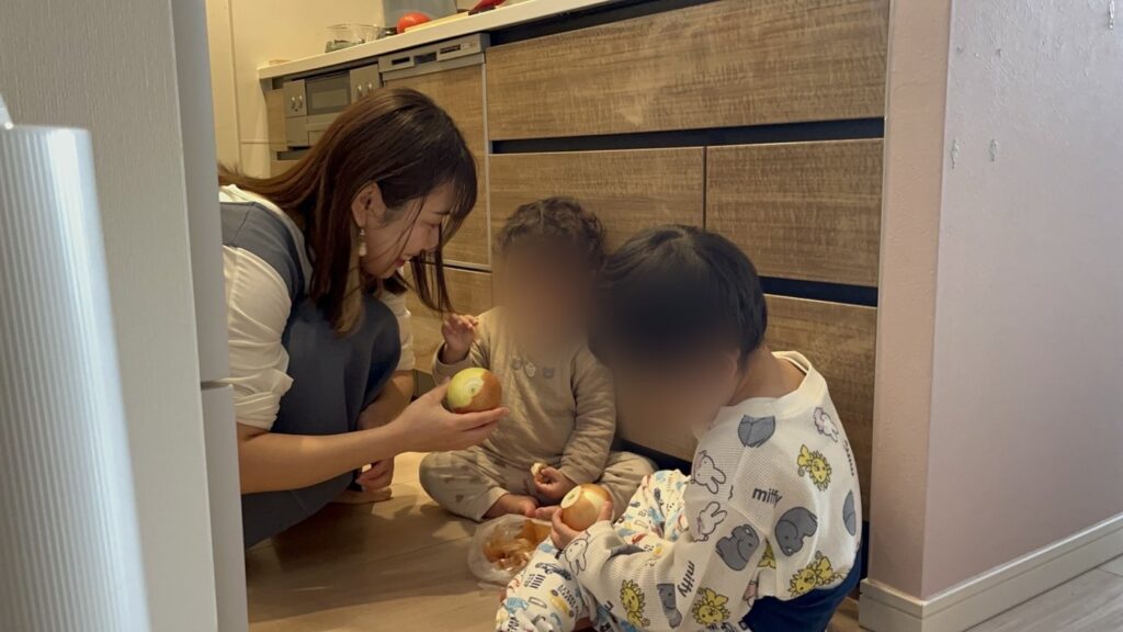 台所の床で、玉ねぎをむく2人の子どもと、声をかけるなかのかほ（お子さんの顔はぼかしています）