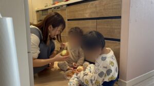 台所の床で、玉ねぎをむく2人の子どもと、声をかけるなかのかほ（お子さんの顔はぼかしています）